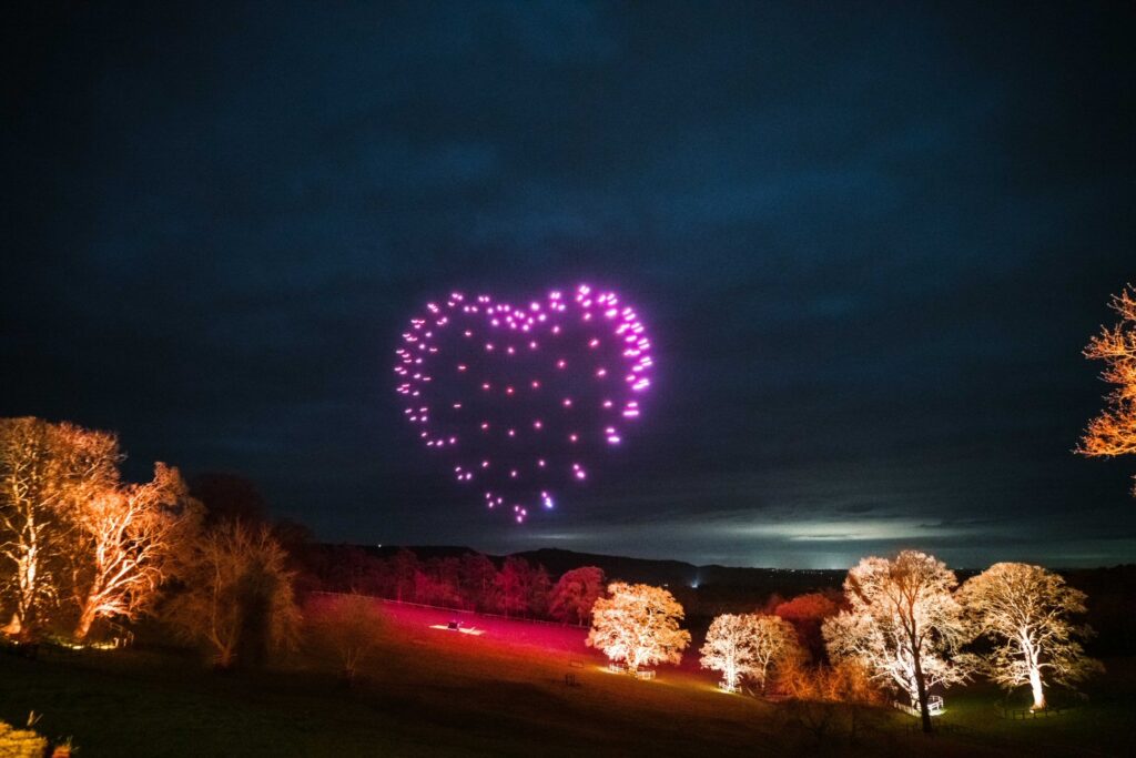 Purple drones form a glowing heart shape in the night sky above illuminated trees in a field.