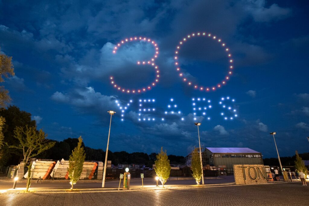 Drones form "30 YEARS" in lights above an outdoor venue at dusk with trees and buildings below.