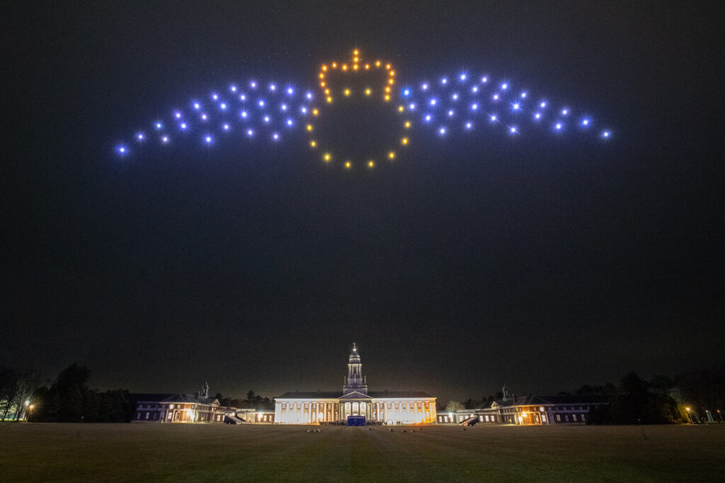 Drones form a glowing crown and wings pattern in the night sky above a large illuminated building.