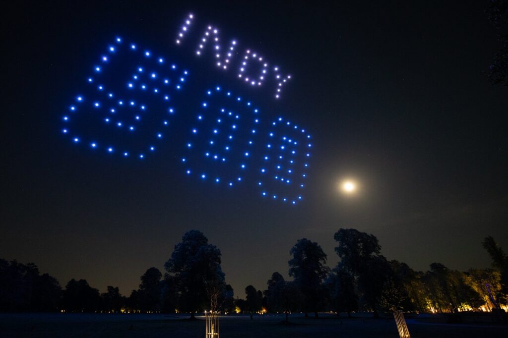 Drones form "INDY 500" in blue lights in the night sky above trees, with the moon visible.