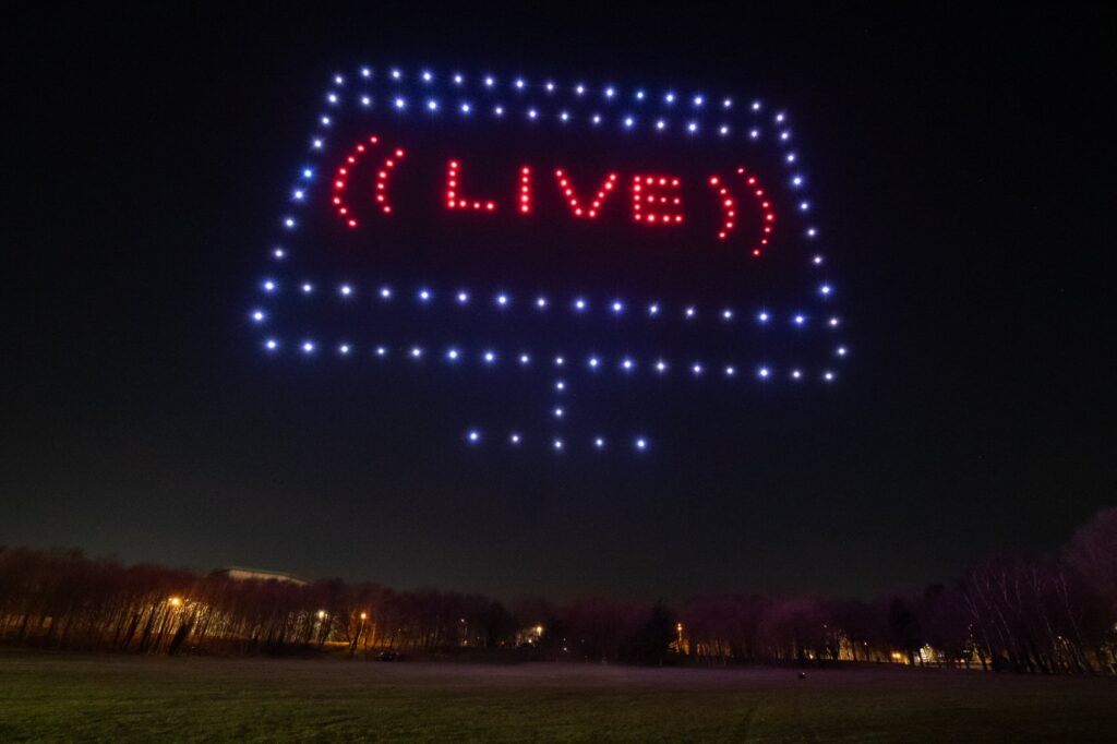 A drone light display forms a screen with the word "LIVE" in red, hovering above a park at night.