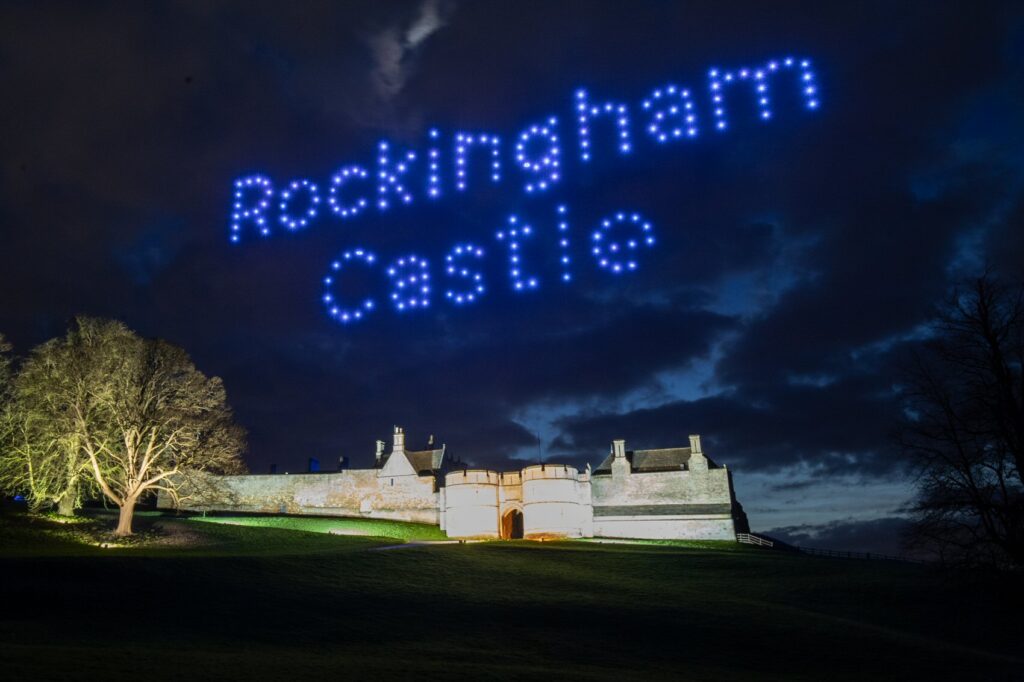 Drones spell “Rockingham Castle” in blue lights above an illuminated castle at night.